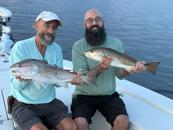 Two men on a boat hold up large redfish. Water and sky in background.