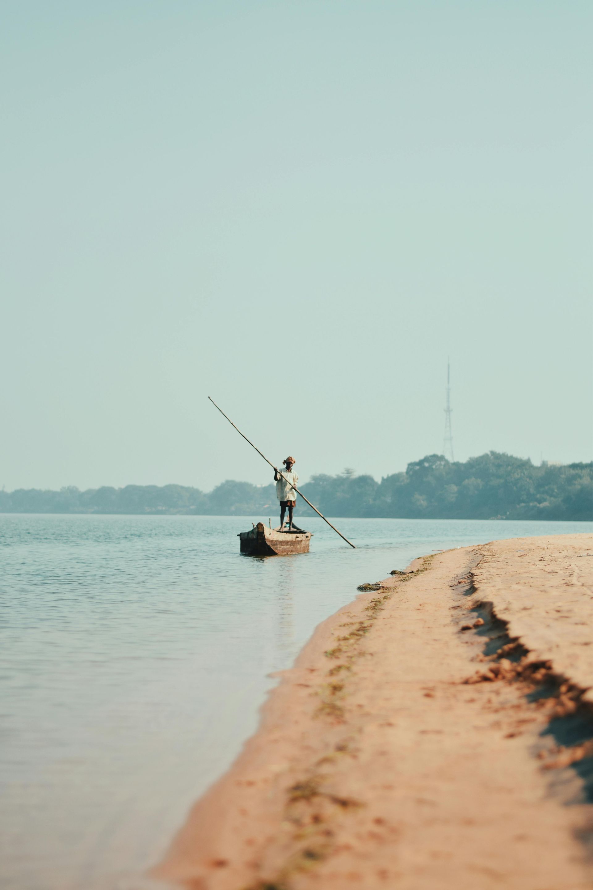 Man in boat with long pole on calm water near sandy shore.