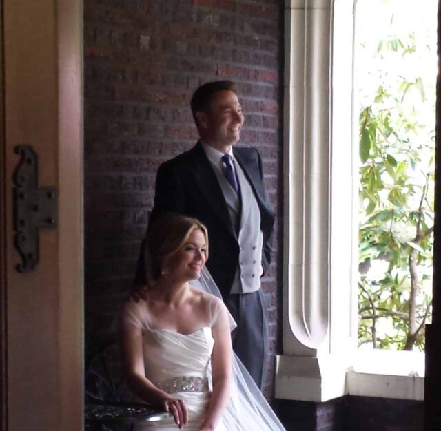 A bride and groom pose for a picture in front of a window