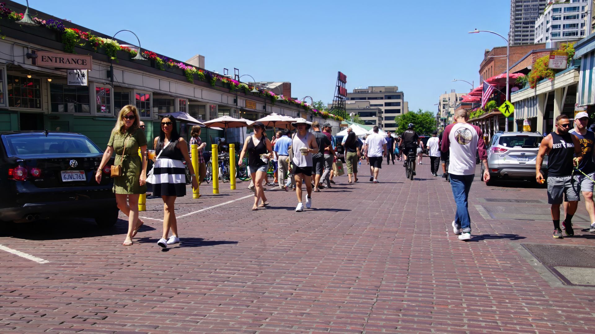 People walking on a brick street in a marketplace with shops and umbrellas on a sunny day.
