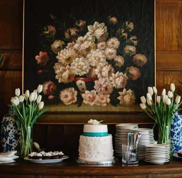 A table with a cake and vases of flowers in front of a painting of flowers