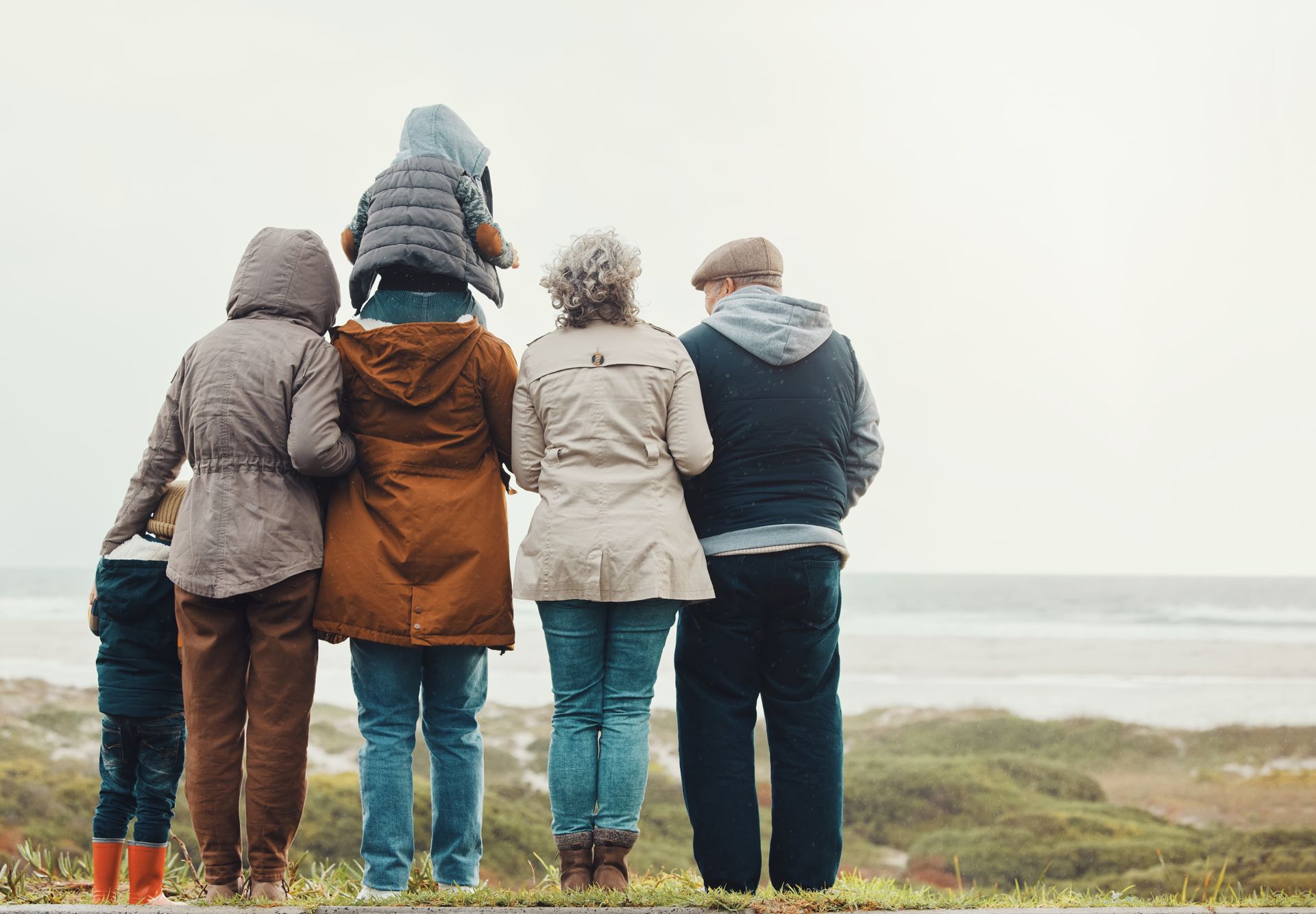 A group of people wearing jackets and coats standing in a line at the beach, looking out toward the water.