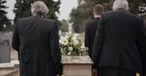 Three people in dark suits carry a casket adorned with white flowers through a cemetery.