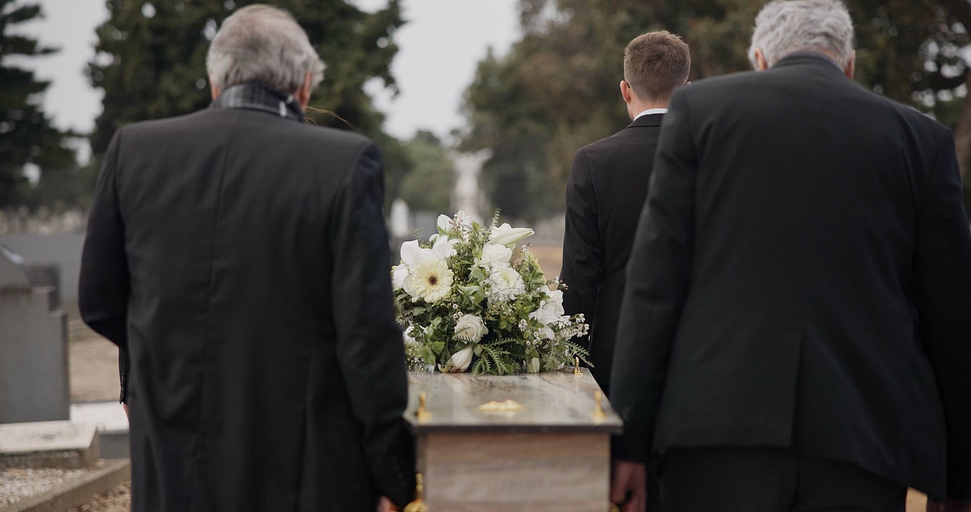 Three people in dark suits carry a casket adorned with white flowers through a cemetery.