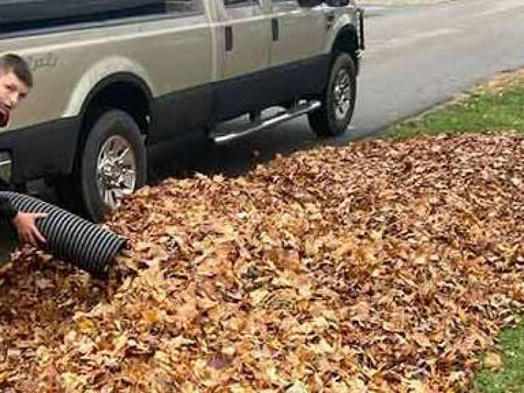 A person uses a black, corrugated hose to vacuum dry brown leaves next to a tan and black pickup truck.