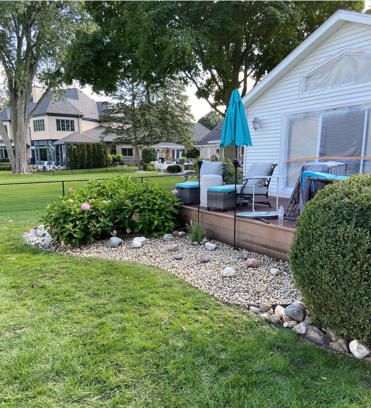 A backyard deck with a blue umbrella, outdoor seating, and a pebble garden bed next to a white house.