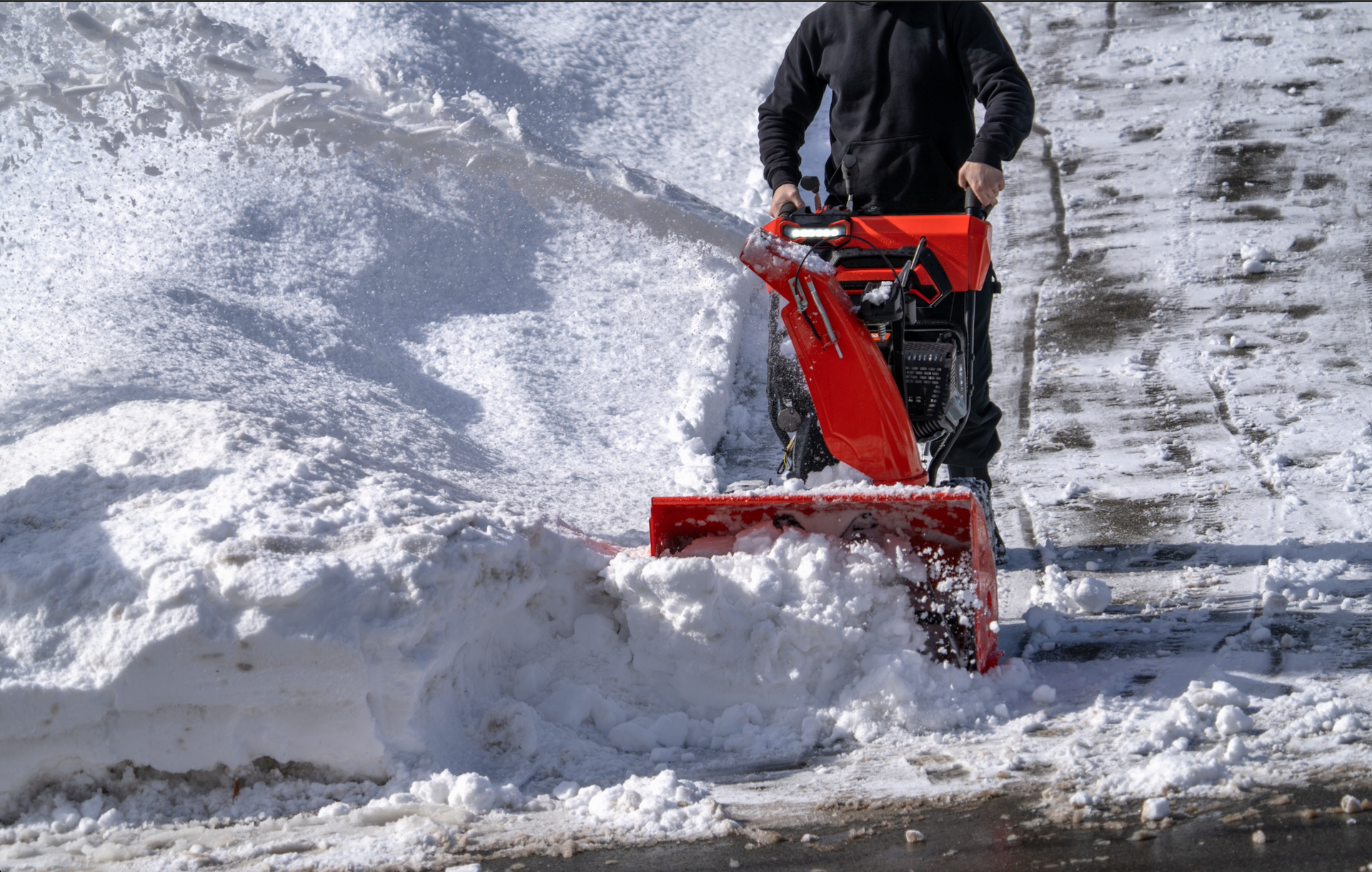A person operates a bright red snow blower to clear a path through deep snow on a sunny winter day.