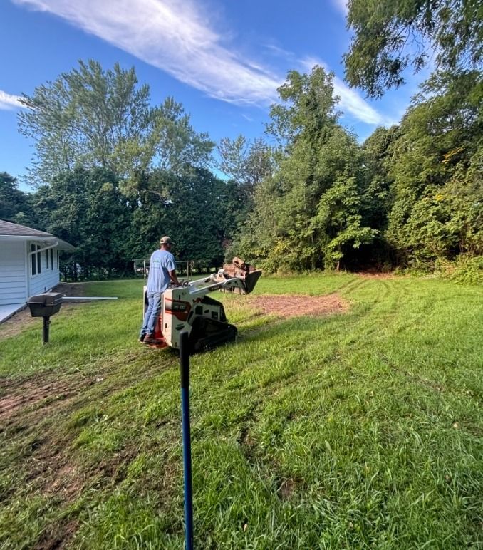 A person operating a white skid-steer loader to clear a patch of dirt in a grassy residential yard on a sunny day.