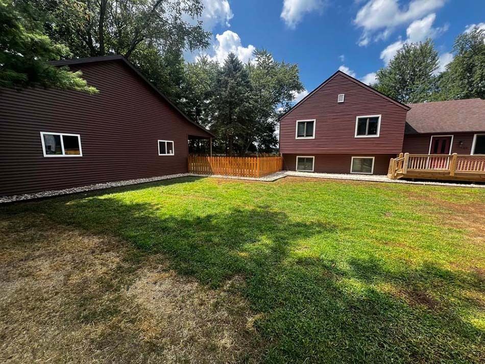 Two maroon houses with white-trimmed windows face a green lawn under a partly cloudy blue sky.
