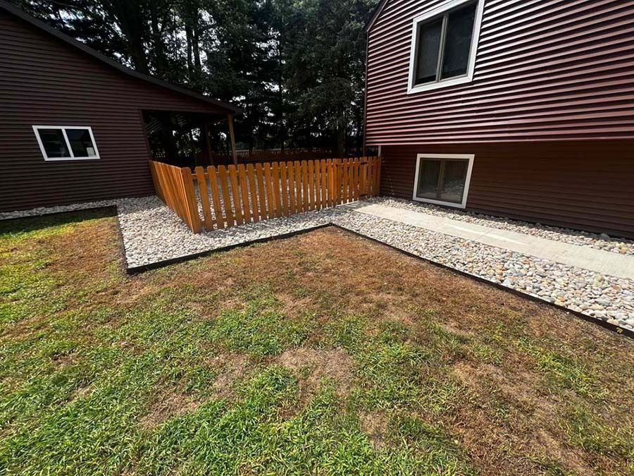 A wooden fence separates a gravel-bordered backyard from a brown house with a paved stone walkway.