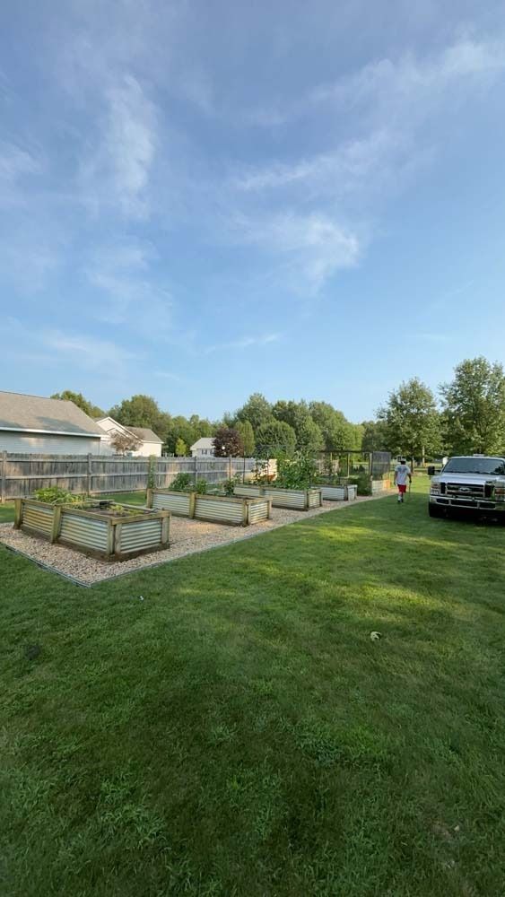 A row of raised garden beds sits on a gravel base in a grassy backyard near a parked vehicle on a sunny day.