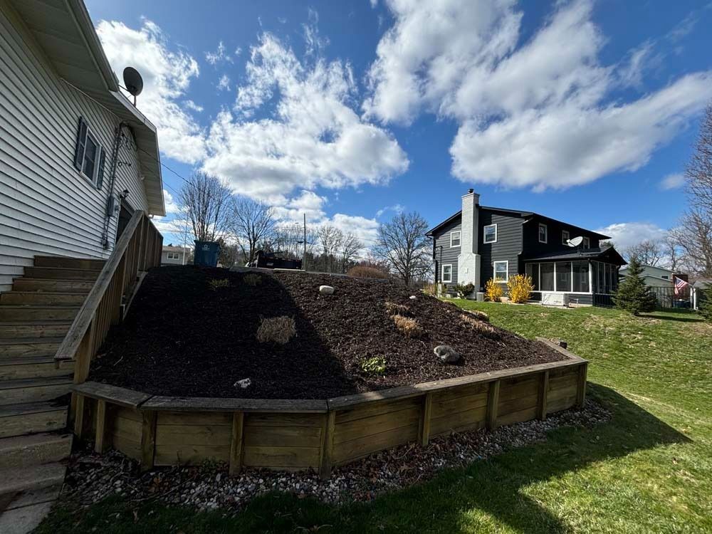 A wooden retaining wall borders a mulched garden bed beside a house, with another house visible in the background.