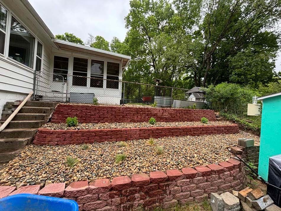 A tiered, reddish-brick retaining wall landscape with gravel beds and small plants next to a white house.