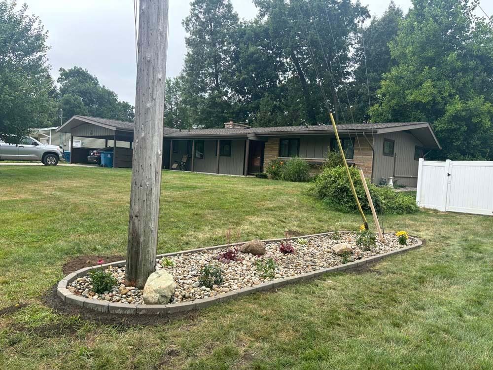 A landscaped garden bed with rocks surrounds a utility pole in a residential lawn, with a one-story house in the background.