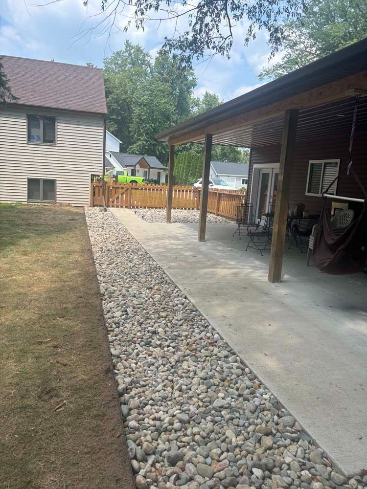 A backyard view of a concrete patio covered by a roof, adjacent to a wide gravel border and a tan house with a wood fence.