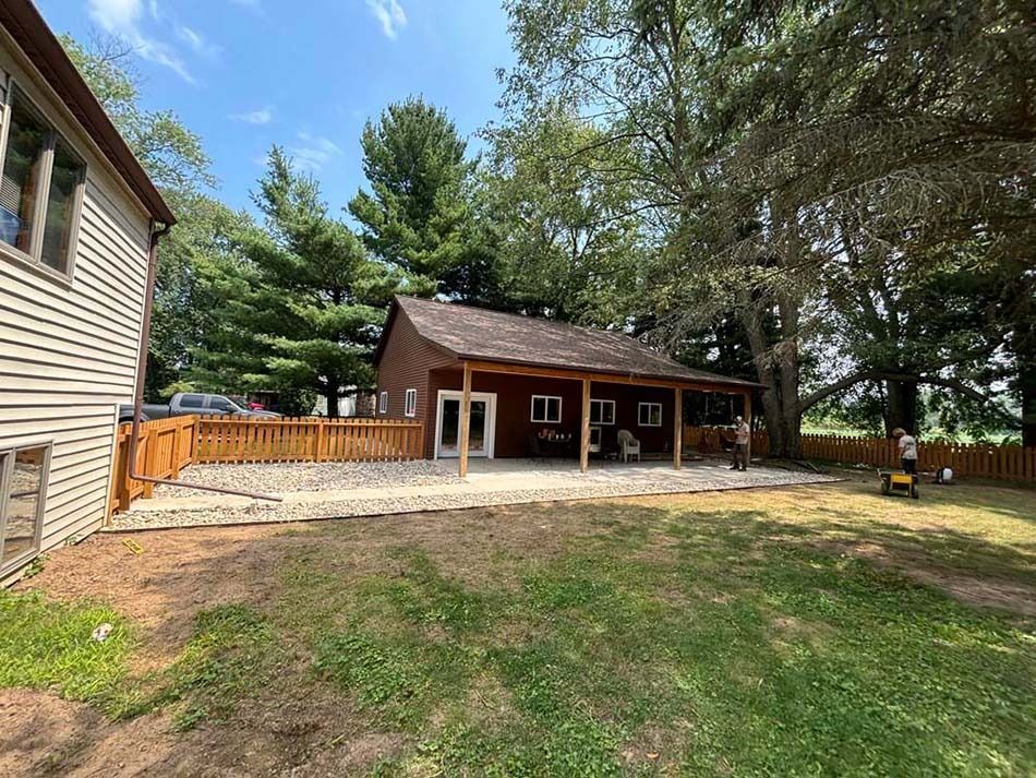 A brown house with a covered patio, a gravel yard, and a wooden fence, set against a backdrop of trees under a blue sky.