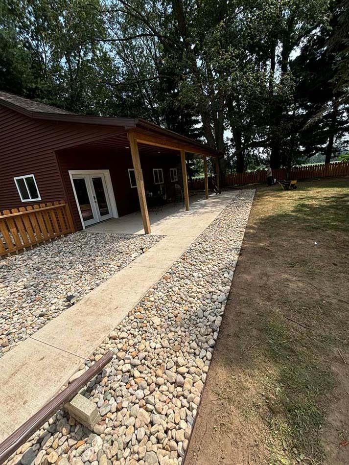 A brown building with a covered concrete porch, surrounded by a border of large river stones, next to a grassy lawn.