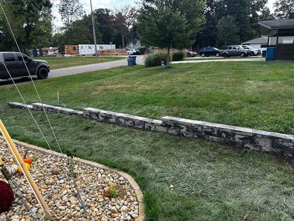 A stone retaining wall sits in a grassy front yard, near a gravel garden bed and a parked truck.