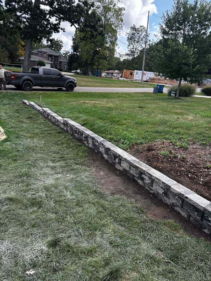 A low, gray stone retaining wall under construction in a grassy residential yard with a truck parked in the background.