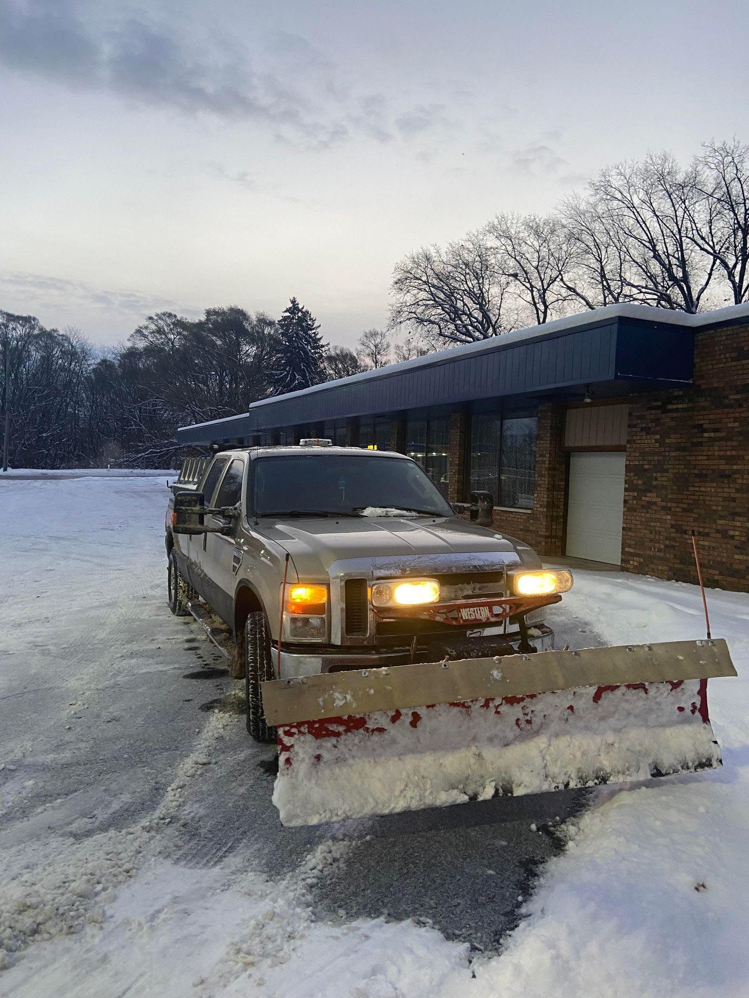 A light-colored pickup truck with a large snow plow attached to the front sits in a snow-covered parking lot.
