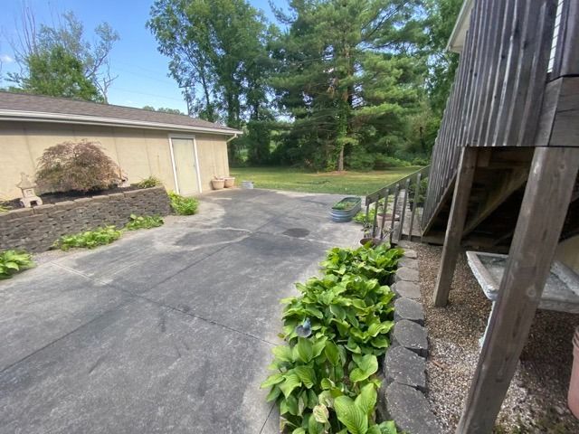 A concrete driveway leads toward a garage and trees, with a stone-bordered garden bed featuring green hostas in the foreground.