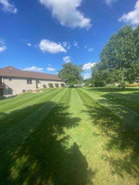 A large, freshly mowed lawn with prominent striping patterns, next to a house under a bright blue sky with scattered clouds.