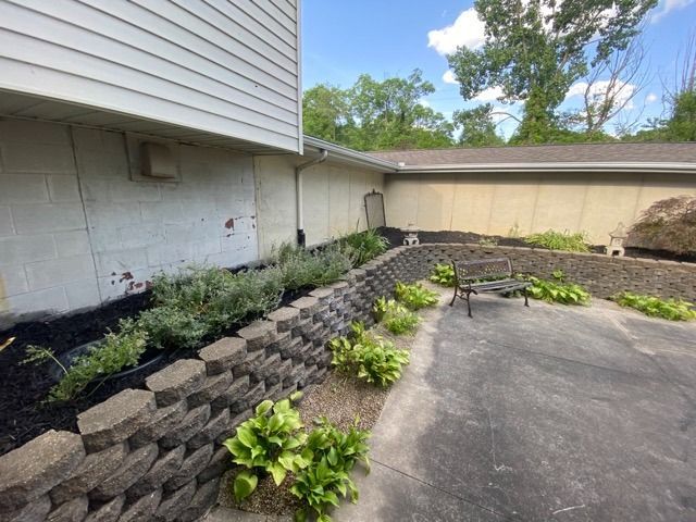 A stone retaining wall curves along a paved area next to a house, with landscaped plants and a small metal bench nearby.