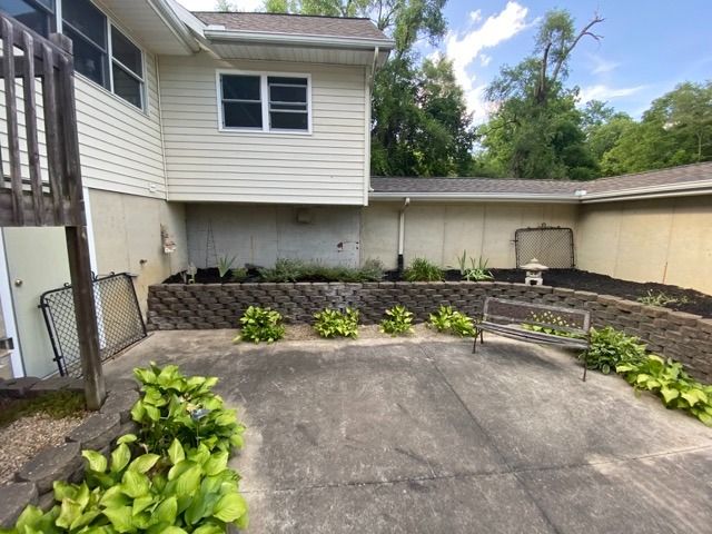 A concrete patio area featuring a low stone retaining wall, green plants, and a small metal bench against a house.