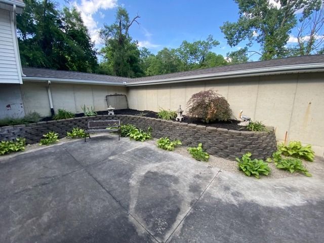A stone retaining wall curves around a corner of a building, framing a small garden with a central Japanese maple tree.