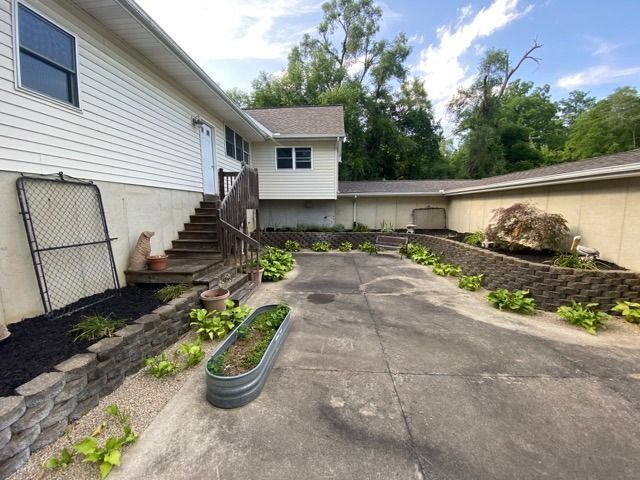 A paved driveway leading to a house with white siding, wood stairs, garden beds, and a stone retaining wall.