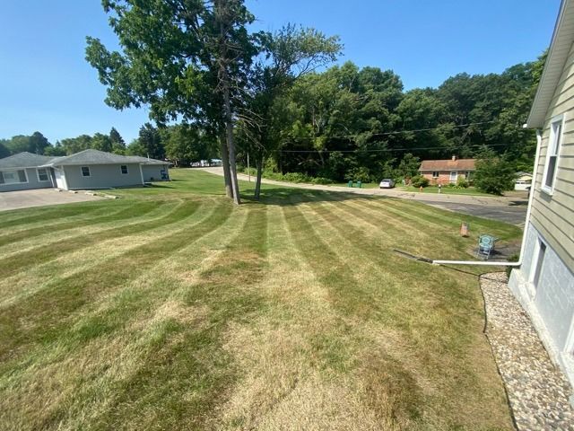 A view of a mowed lawn with alternating dark and light green striped patterns, leading toward trees and a neighboring house.