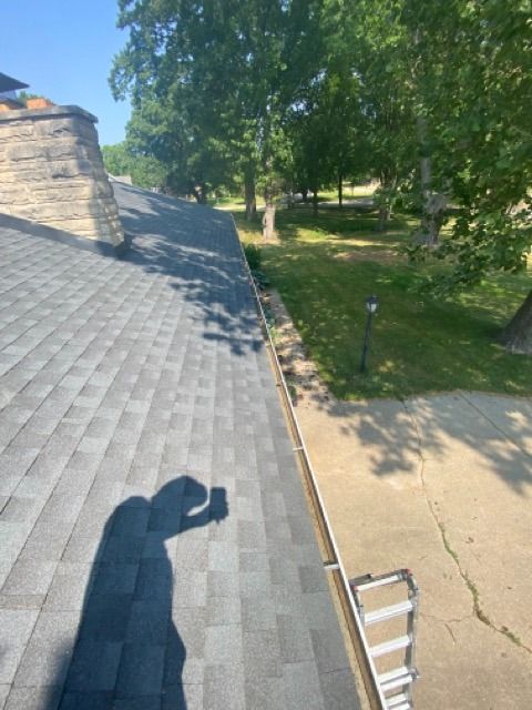 A first-person view from a shingled roof looking down at a concrete driveway, a grass lawn, and a ladder leaning against.