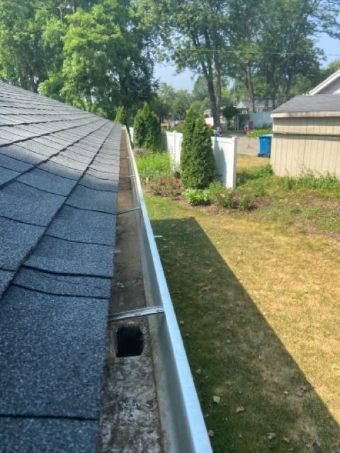 A view looking down a roofline into a metal gutter with a rectangular drainage hole in a residential yard.