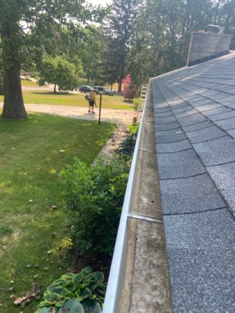A view from a roof showing a gutter next to shingled roofing, with a person standing on the lawn below.