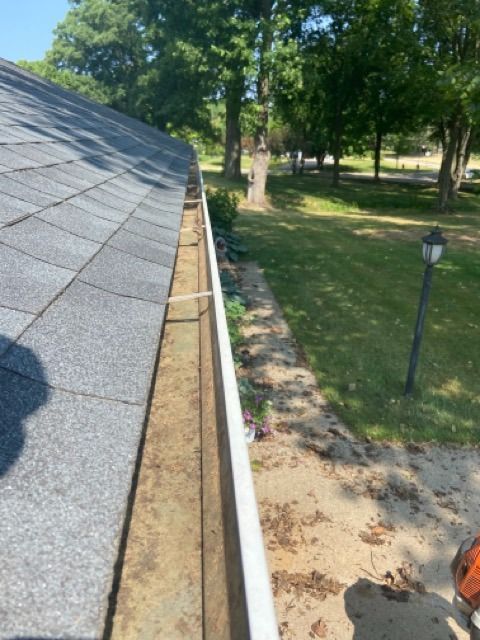 An elevated, high-angle view showing a dirty residential roof gutter filled with debris, with a yard visible below.