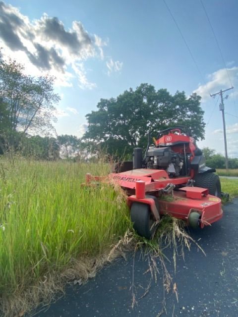 A red zero-turn mower positioned at the edge of a paved road, beginning to cut a field of tall grass under a blue sky.