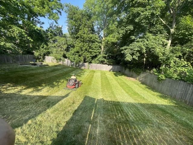 A person riding a red lawn mower cuts grass in a large, sunny backyard surrounded by trees and a wooden fence.