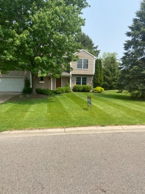 A two-story house with tan siding and stone accents sits behind a freshly mown lawn with stripes under a clear blue sky.