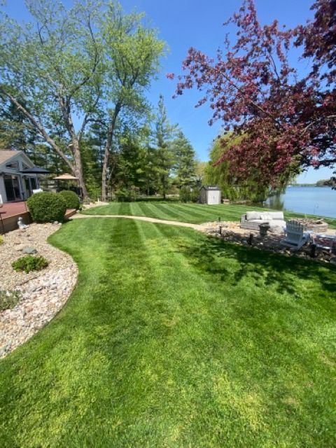 A manicured lawn with stripe patterns leads toward a lake, surrounded by trees, garden beds, and a patio area.