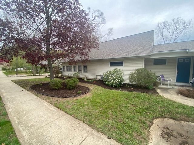 A low-slung, light-colored home with dark trim and a blue front door, featuring a front yard garden and a large tree.