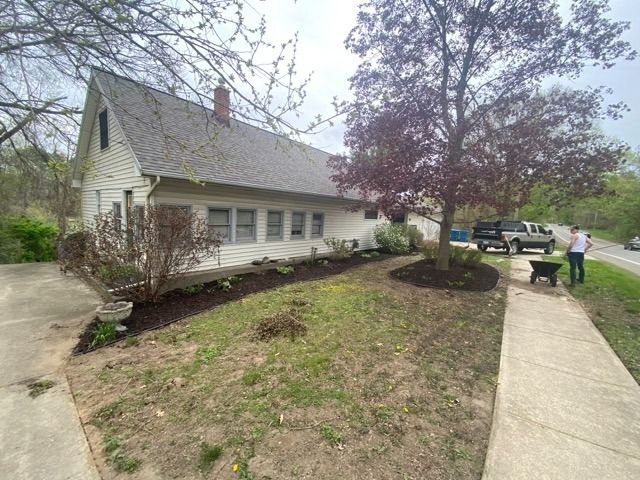 A side view of a white farmhouse with a large tree in the yard and a person working on the landscaping near a sidewalk.
