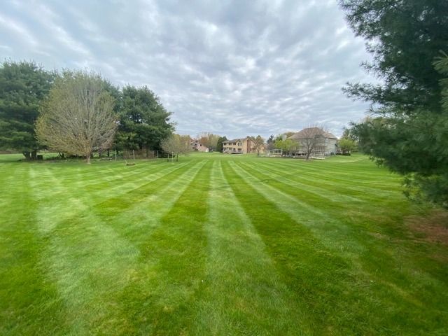 A wide, lush green lawn featuring uniform mowing stripes, leading toward distant houses under a cloudy sky.