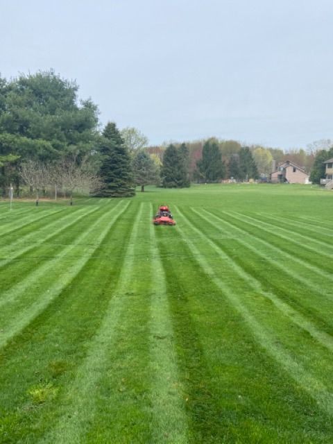 A person rides a lawnmower across a large, open field with freshly mowed, alternating dark and light green grass stripes.