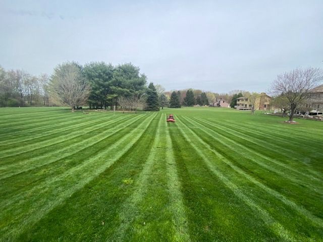A sprawling green lawn with freshly mowed stripes leading toward a distant red riding mower and trees under a cloudy sky.