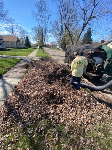 A person in a yellow long-sleeved shirt uses a vacuum hose to clear a large pile of dry leaves on a suburban street.