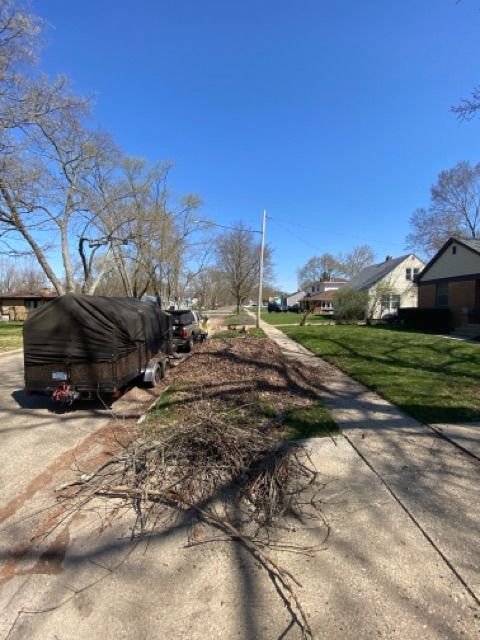 A covered trailer parked on a residential street next to a large pile of branches on the sidewalk.