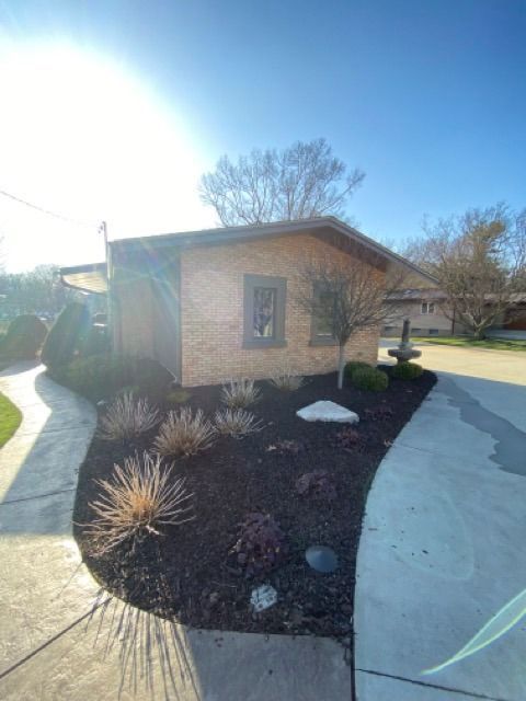 A tan brick house with a dark roof surrounded by dark mulch, landscaping, and concrete walkways under a clear blue sky.