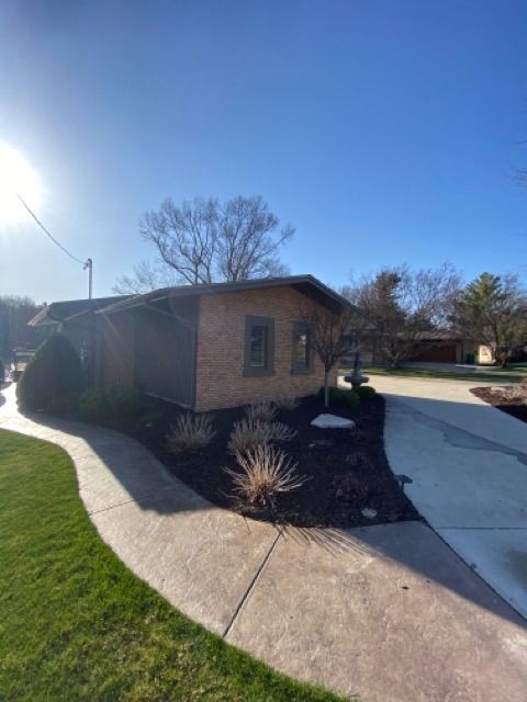 A tan brick house with a dark roof on a sunny day, featuring a concrete walkway, mulch garden beds, and a driveway.