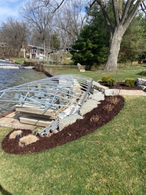 A metal boat lift frame sits on the shore next to a landscaped mulch bed and large stone boulders by a lake.