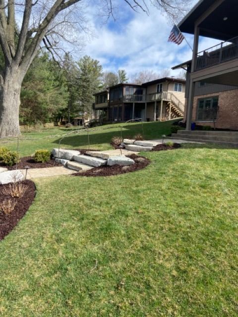 A sloped grassy lawn featuring tiered stone steps and mulch beds leading toward two multi-story houses.